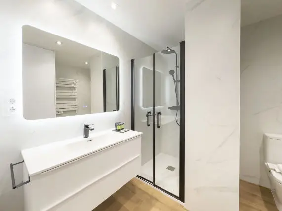 vista parcial donde aparece la ducha al fondo junto con el inodoro en la imagen se observa parte del lavabo con mueble bajero apartamento de lujo Alfisa B2